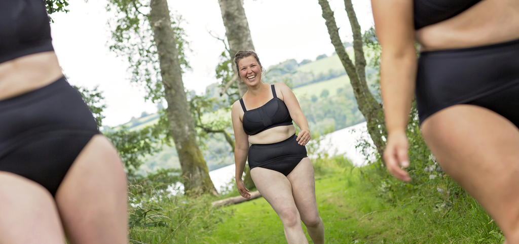 Three models are wearing the Black Molke Swim sets and walking towards you in the woods with the loch behind them through the trees. The two on the left and right are really close to the camera and out of focus you can only see their torsos and tops of legs. The model in the middle is further back and in focus She is smiling widely.