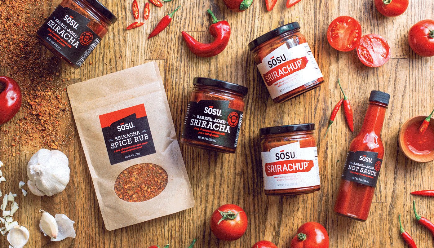 Various Sosu Sauces products including spice rubs, barrel-aged sriracha in jars, hot sauce, and ketchup spread out on a tabletop with garlic cloves, tomatoes, peppers, and spices throughout.