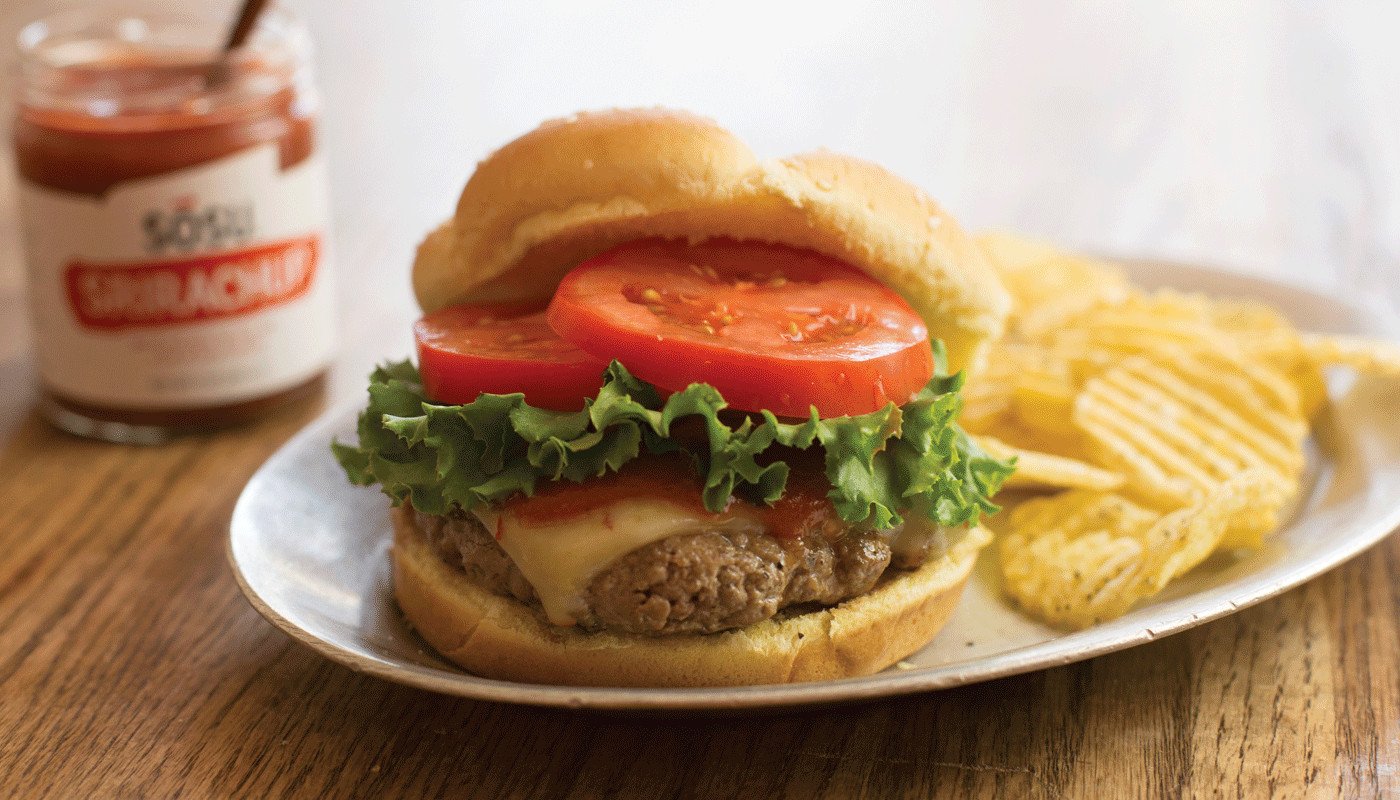 A cheeseburger sitting on a plate with potato chips and a jar of Sosu Sauces Sriracha-flavored ketchup in the background.