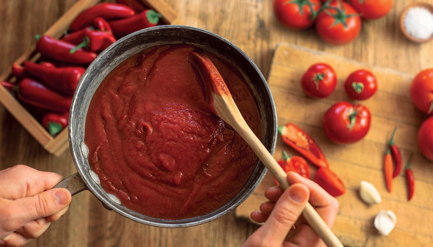 A person holding a saucepan filled with sauce and stirring with a wooden spoon. Peppers, tomatoes, and garlic are in the background.