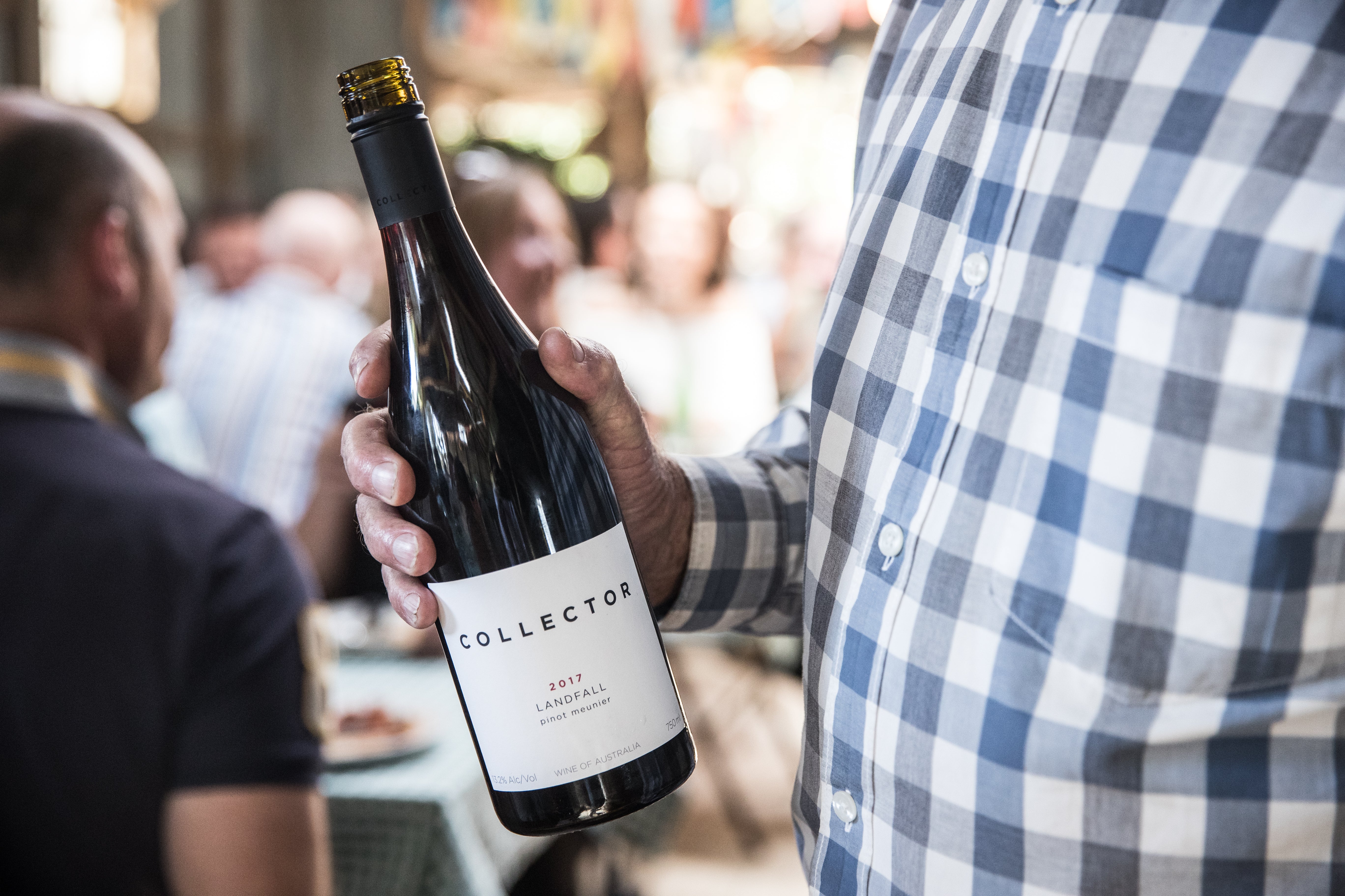 A gentleman in a blue checked shirt holds a bottle of the Landfall Pinot Meunier. In the background, people are celebrating.