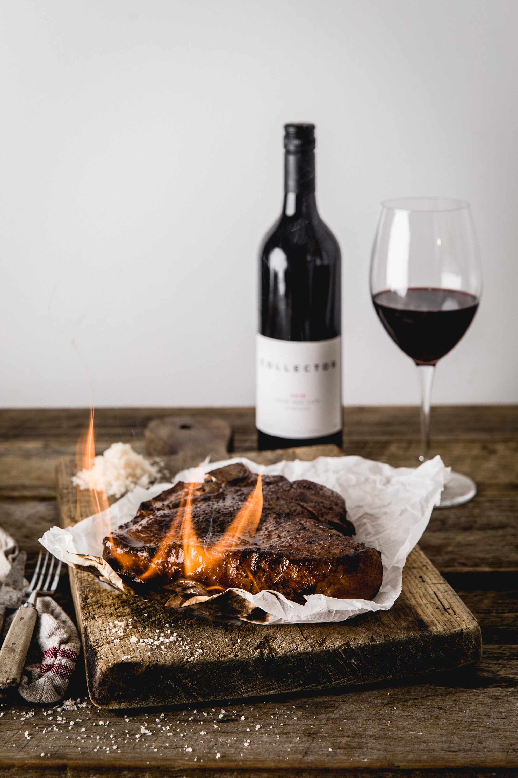 A T-bone steak, grilled and with flames still licking its sides, rests on a rustic wooden board. A bottle and glass of Collector Wines Rose Red City Sangiovese sits in the background.