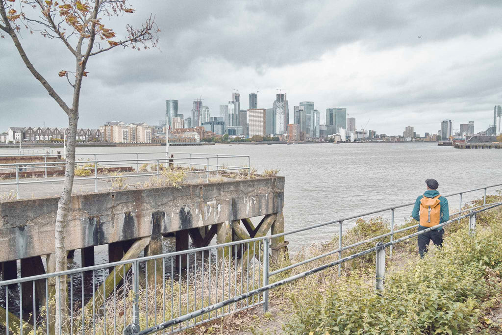 Photographer Joe McGorty wears an orange waterproof backpack looking across the Thames to the London Docklands skyline