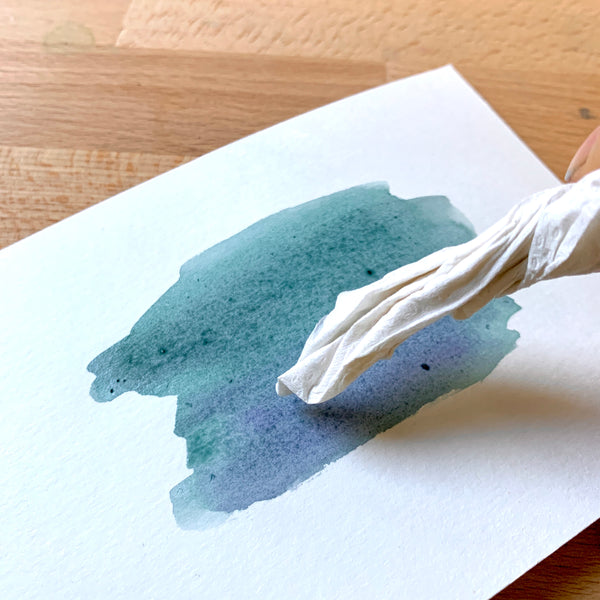 Close up of a piece of watercolour paper and paint, with paper towel dabbing the paint
