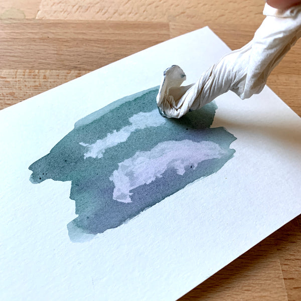 Close up of a piece of watercolour paper and paint, with paper towel dabbing the paint