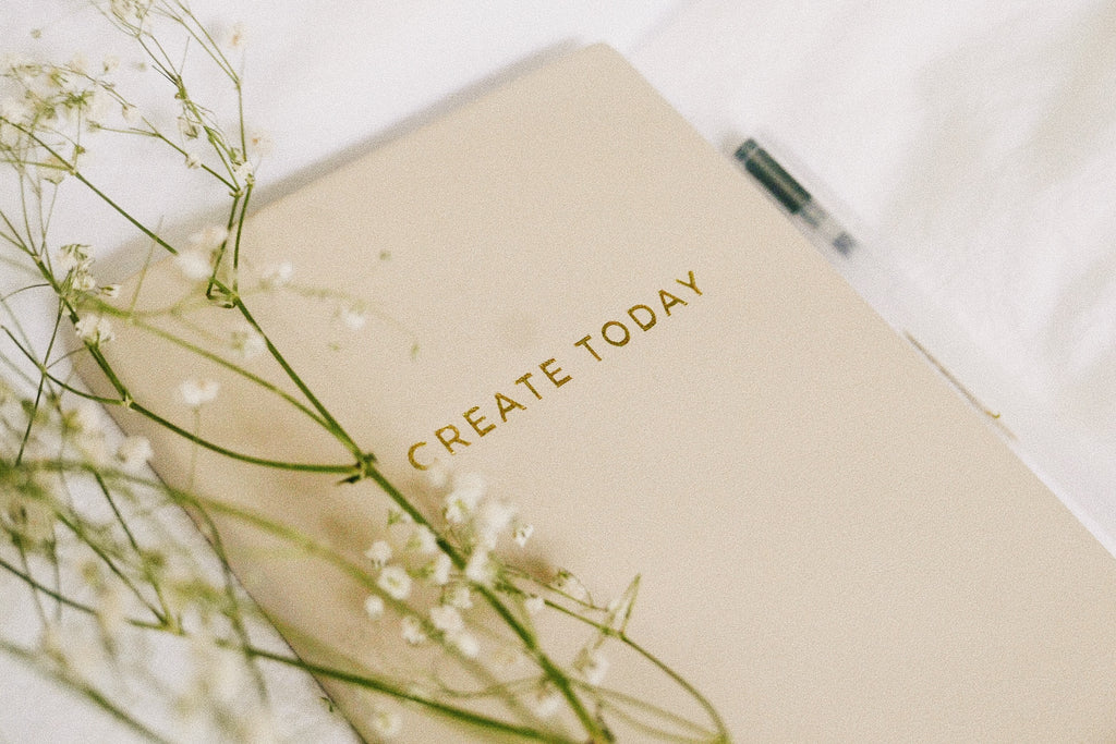 Close up on a pink journal with the words Create Today on it, with some delicate flowers placed over top