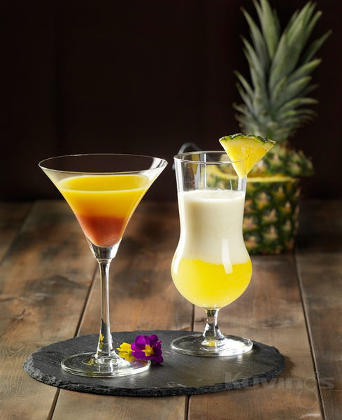 2 cocktails on wood table in front of whole pineapple