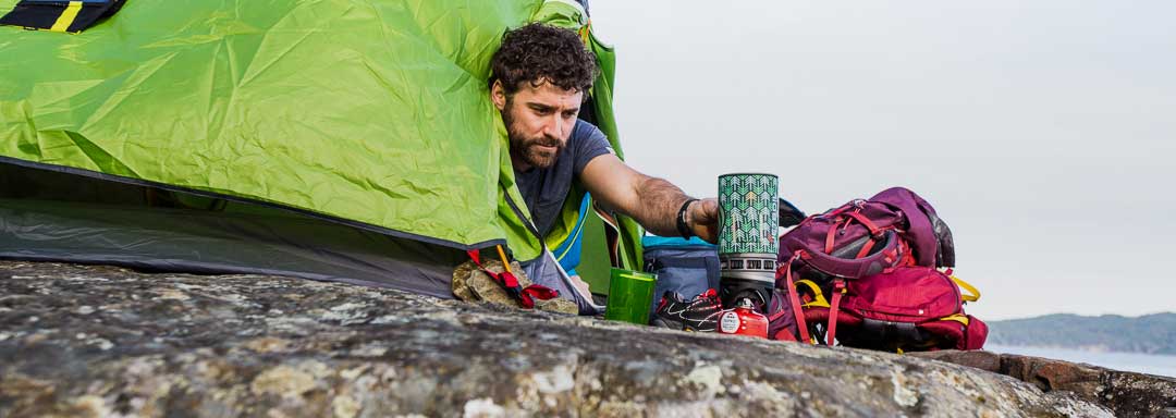 A backpacker reaches out from a bright green tent to tend to a camp stove set up beside backpacks and gear