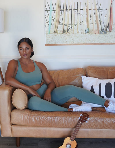 Beautiful Hitraan Model sitting on a couch playing the ukelele and wearing the Crimson tavernorlando Essential Leggings and Leilani Sports bra in the colors Juniper