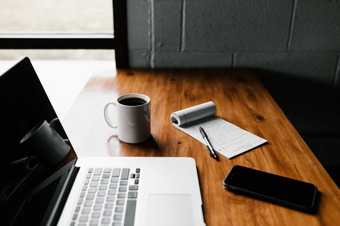 A desk with a coffee, phone, laptop and notepad