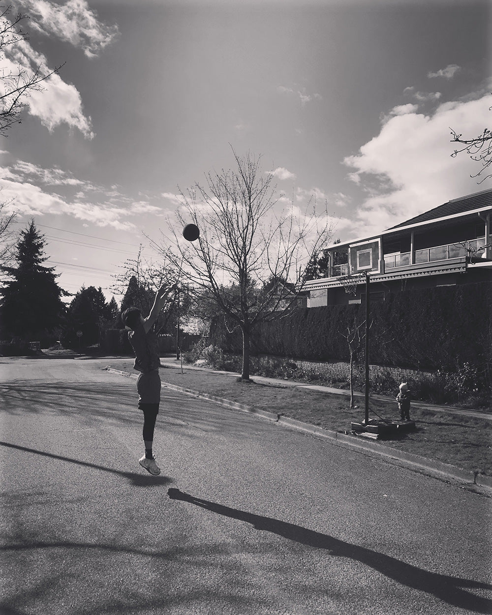 SPITGAN WEBZINE #3 Photo 10. Black and white photo of young man on the street shooting hoops with young toddler watching. Vancouver, BC