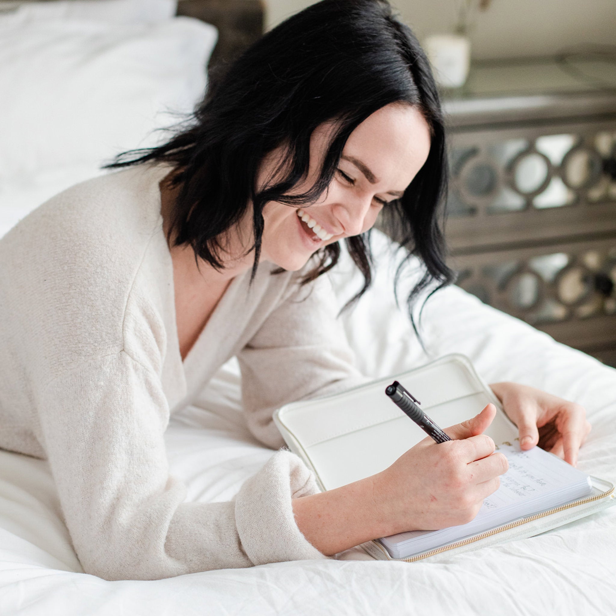 Woman lying on a bed laughing and writing in her journal. 