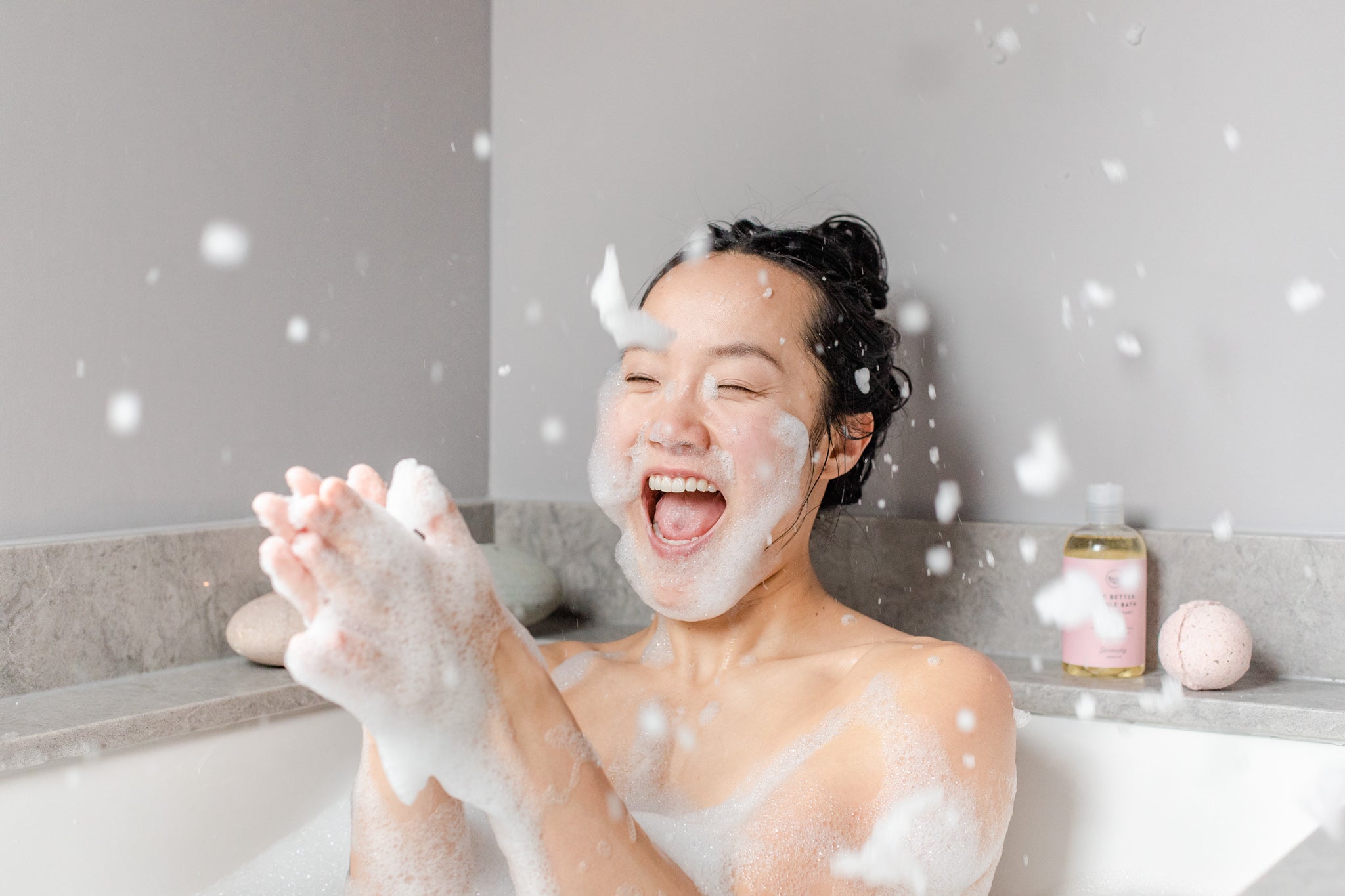 Woman laughing in the bath tub clapping her hands together throwing bubbles in the air with all-natural, local, Canadian SLS-free bubble bath from Rocky Mountain Soap Company.