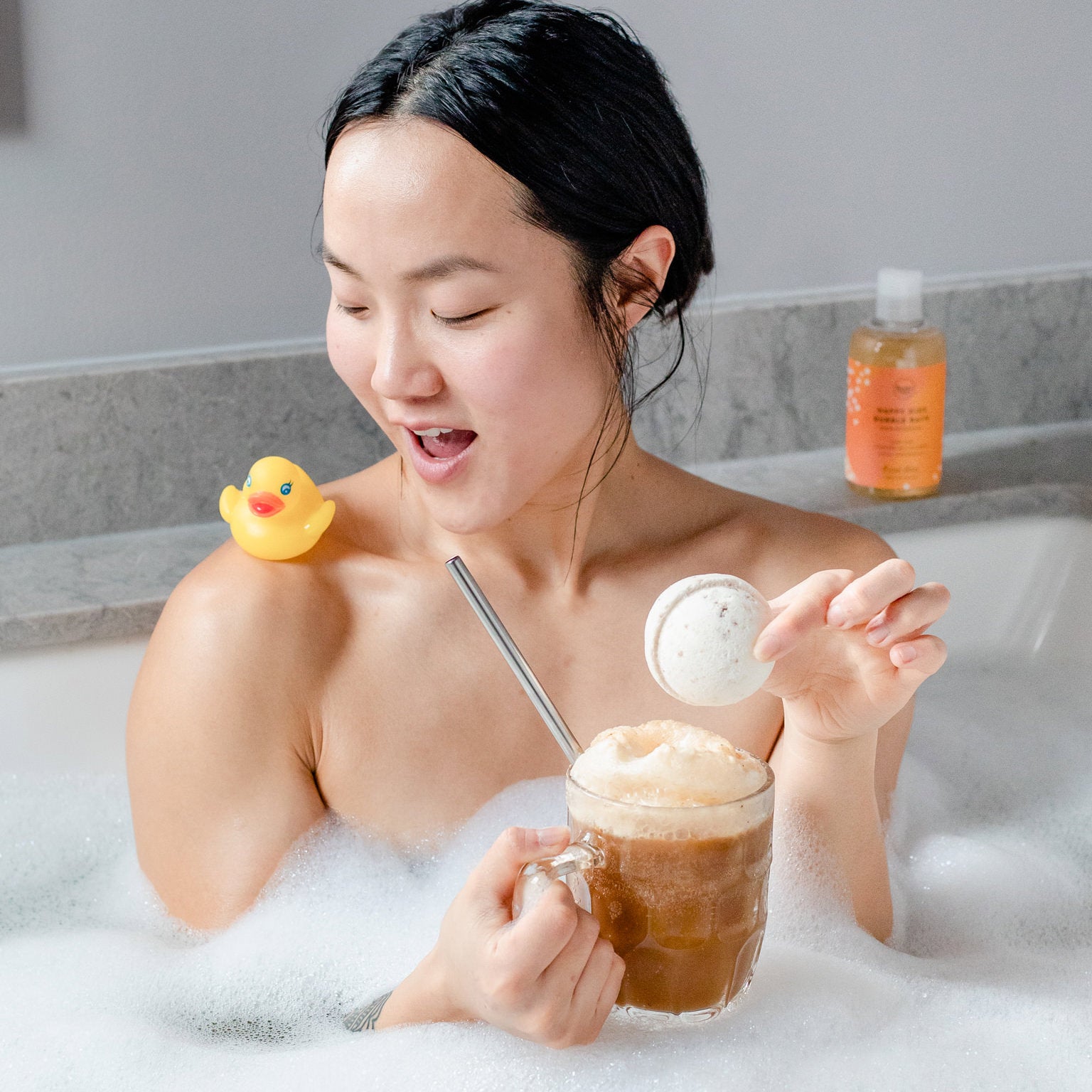 Woman in the bath holding a root beer float with a rubber ducky on her shoulder and an all natural bath bomb in one hand. 