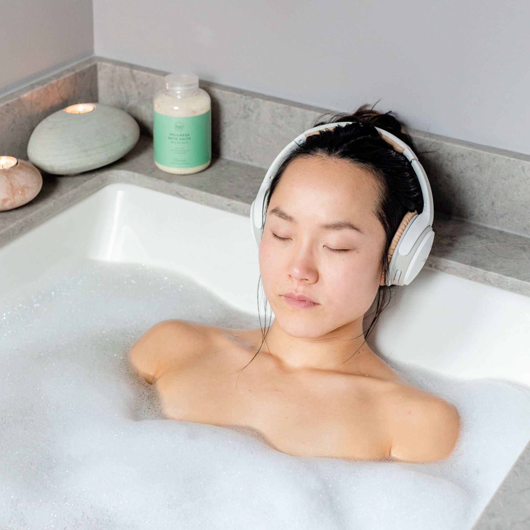 Woman in bath with headphones on, jar of all natural locally made Rocky Mountain Soap Company bath salts in the background. 
