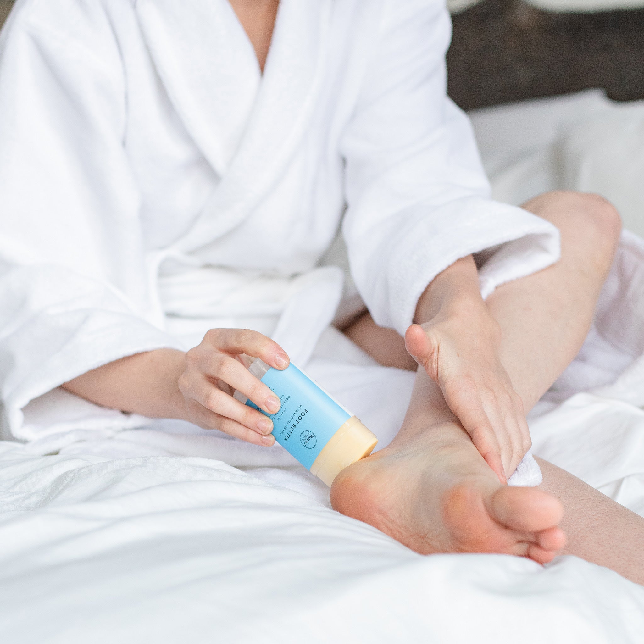 Image of woman in bath robe rubbing Foot Butter on her heel. 