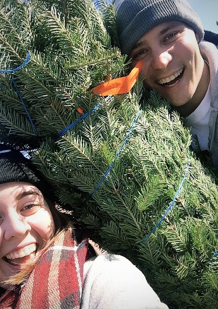 Carina and her partner holding a Christmas tree smiling at the camera. 