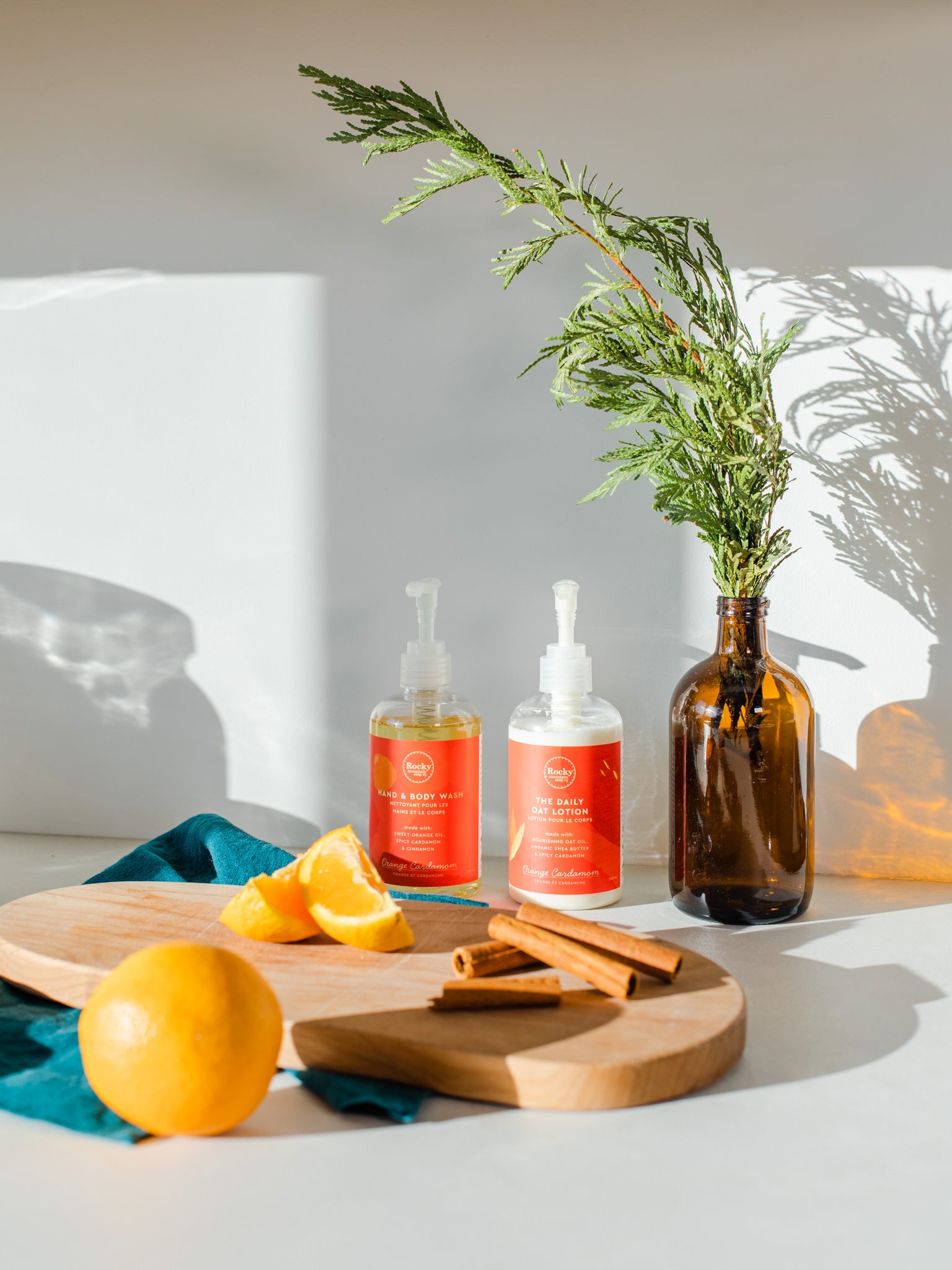 An orange, orange slices, and cinnamon sticks on a kitchen counter next to bottles of Rocky Mountain Soap Company's Orange Cardamom Daily Oat body lotion and liquid hand and body wash.