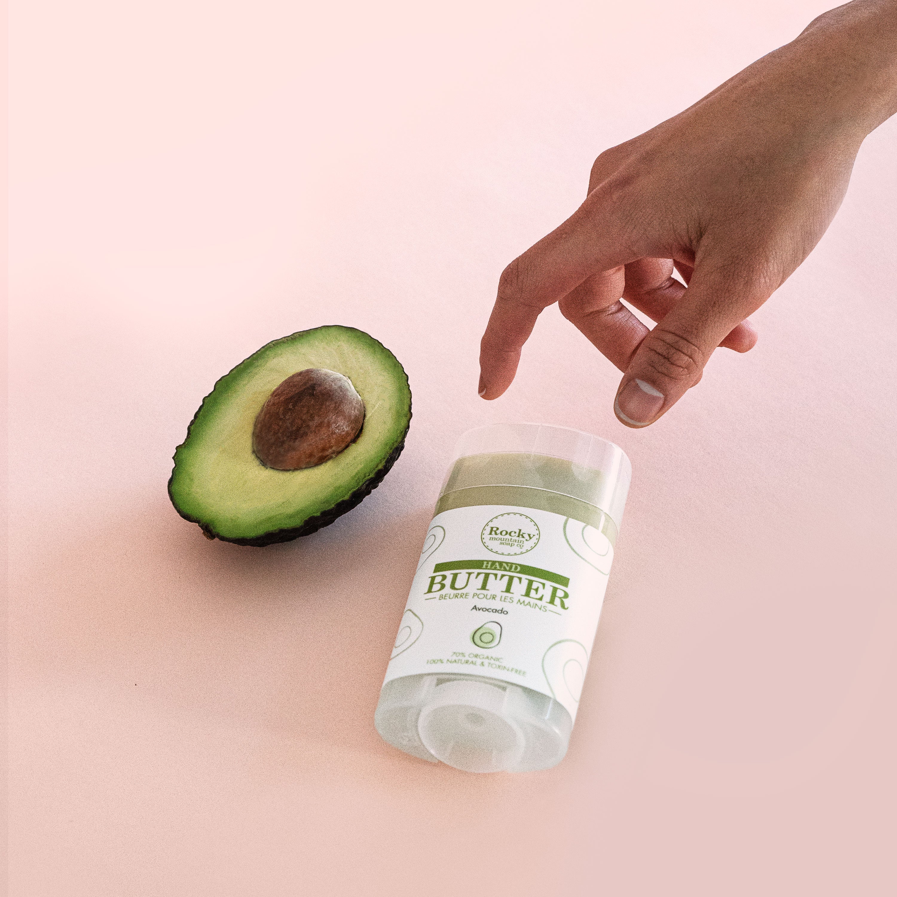 A hand reaches for a Rocky Mountain Soap Company Hand Butter that's place next to half an avocado on a pink background. 