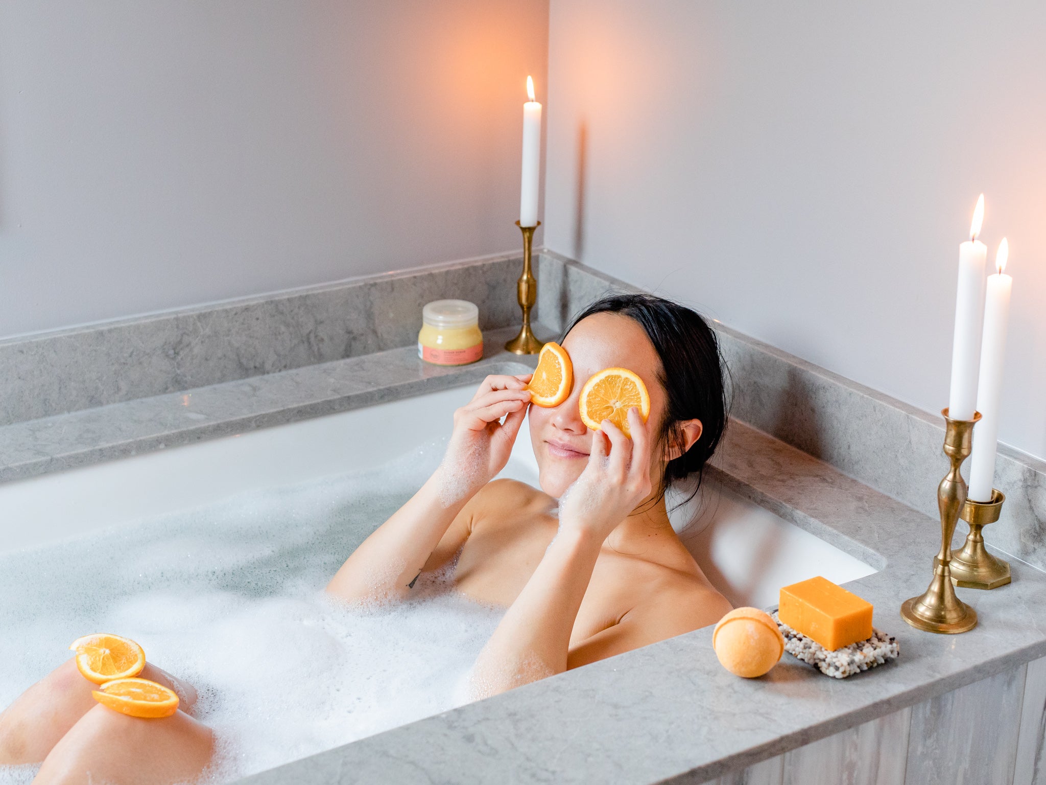 Woman in the bath with candles on the side of the tub and two orange slices over her eyes.