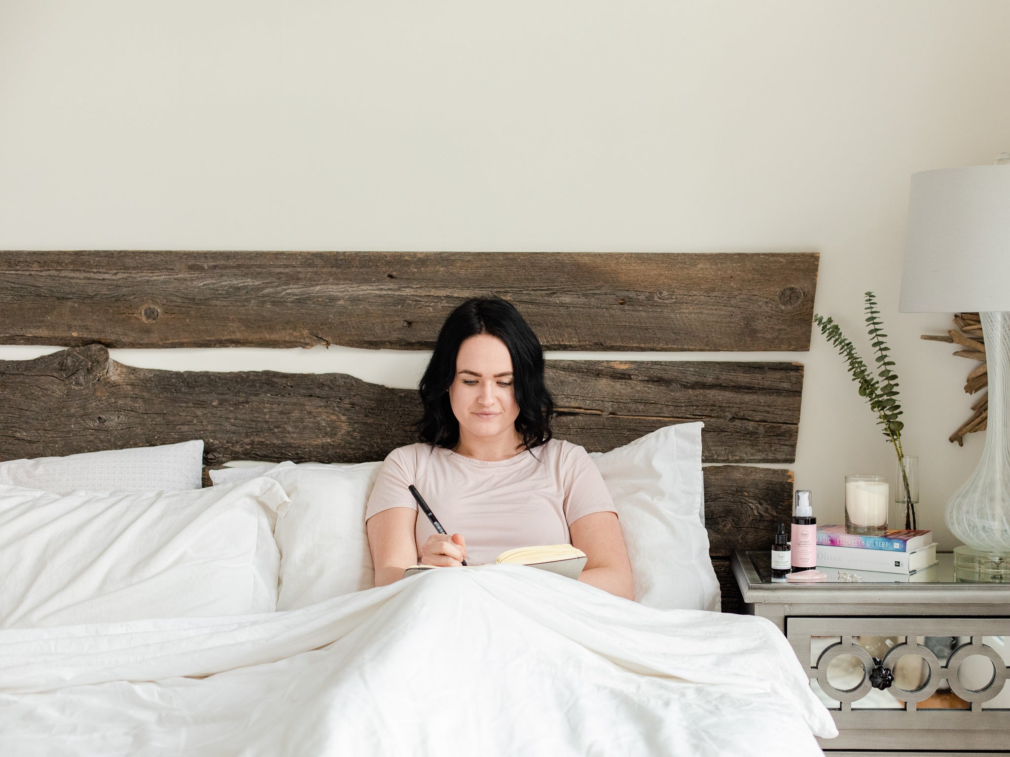 Woman lying in bed writing in a notebook with all natural massage oil from Rocky Mountain Soap Company on her bedside table.