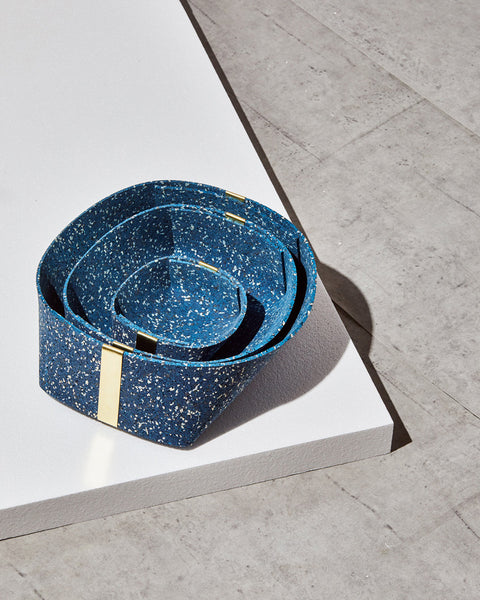 Three speckled blue rubber and brass baskets, nested inside one another on white foam board on concrete surface