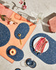 Spring table setting with speckled blue rubber capsule placemats, capsule trivets and brass ring coasters on concrete and terracotta surface. The setting is styled with cut half peach, brass spoon, water pitcher, glass and pink flowers.