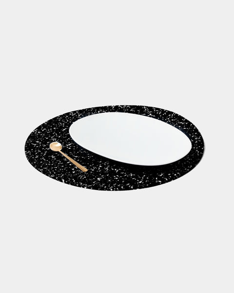 Speckled black round placemat with white place and brass spoon on white surface.