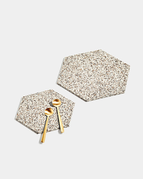 One small and one large hexagon speckled beige rubber trivet on white surface. Small trivet has two brass tea spoons placed on its surface.