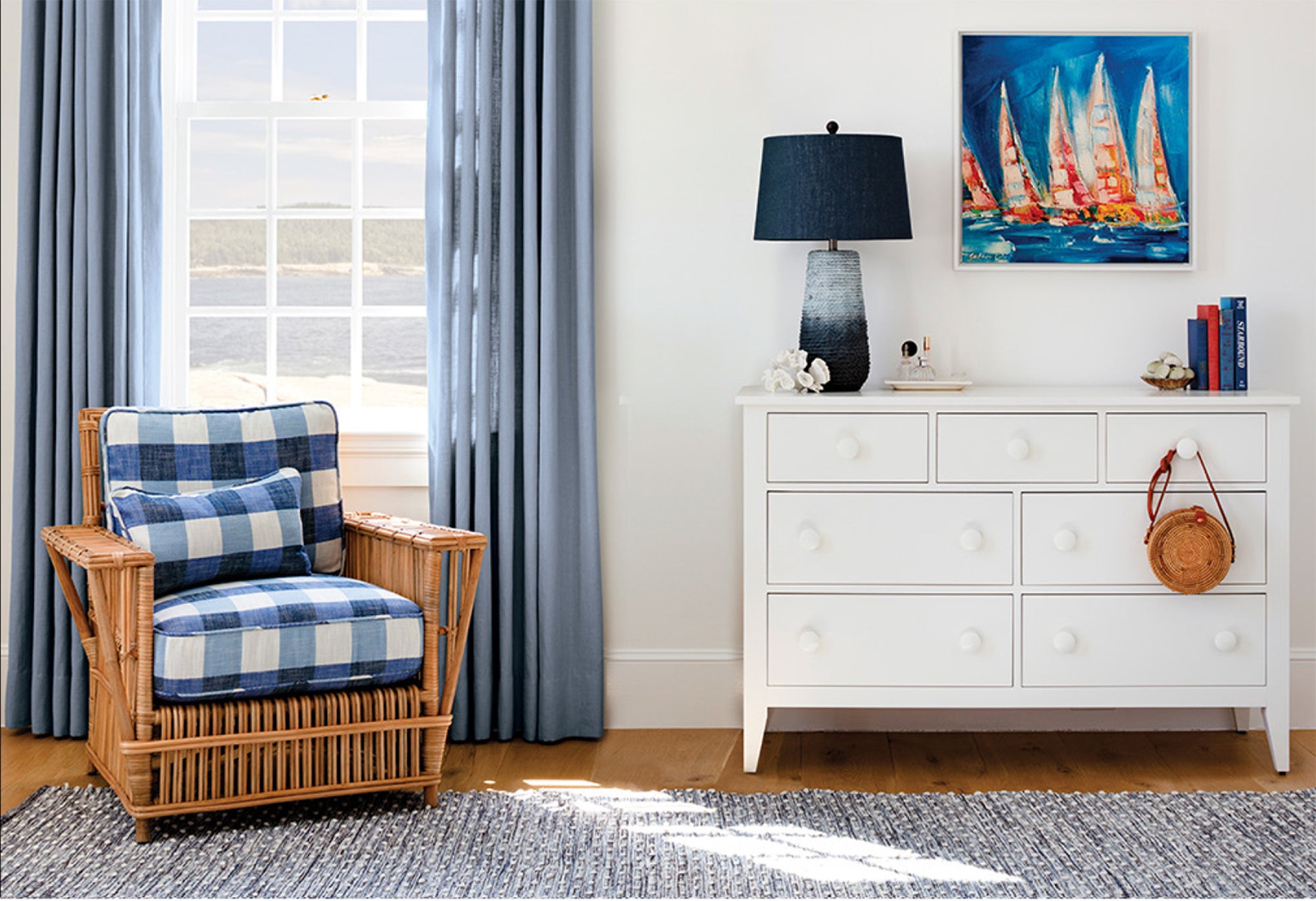 Room with white dresser and blue accents: wicker chair with denim cushions, Marlow lamp, indoor/outdoor rug, Regatta artwork