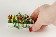 A hand delicately placing a miniature window box with flowers in it onto a white background.