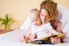 Mother reading to a two year old while both lay on a bed looking at a book.