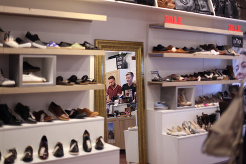 Photo shows inside of the Vegan Style boutique. It shows a mirror which reflects the image of two men, Vegan Style owners Justin Mead and Gavin Reichel. Surrounding the mirror are shelves displaying shoes
