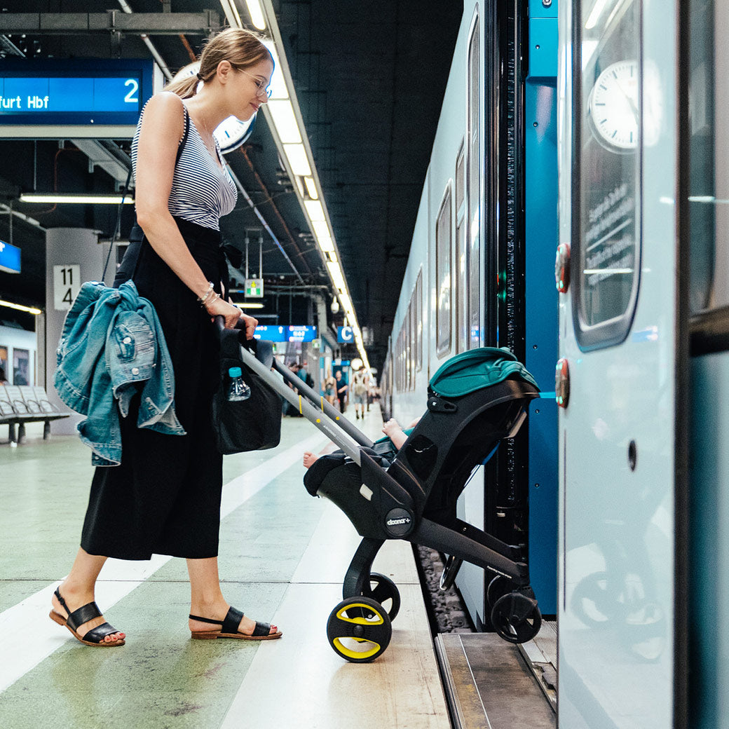 Mother walking onto a train while pushing a baby in the Doona Infant Car Seat and Stroller in -- Color_Racing Green