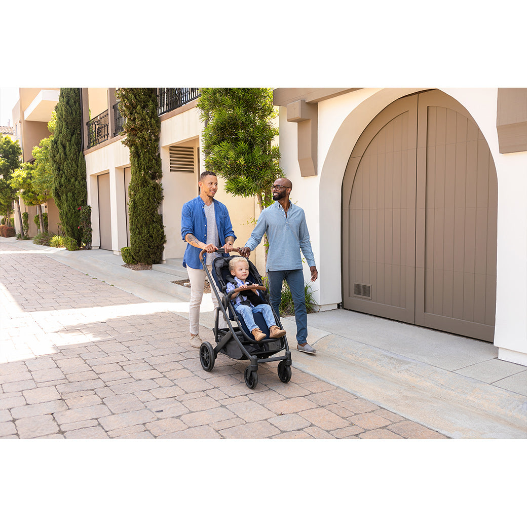 Two dads smiling and pushing a toddler boy with curly blonde hair in a forward facing uppababy cruz v2 stroller -- Lifestyle