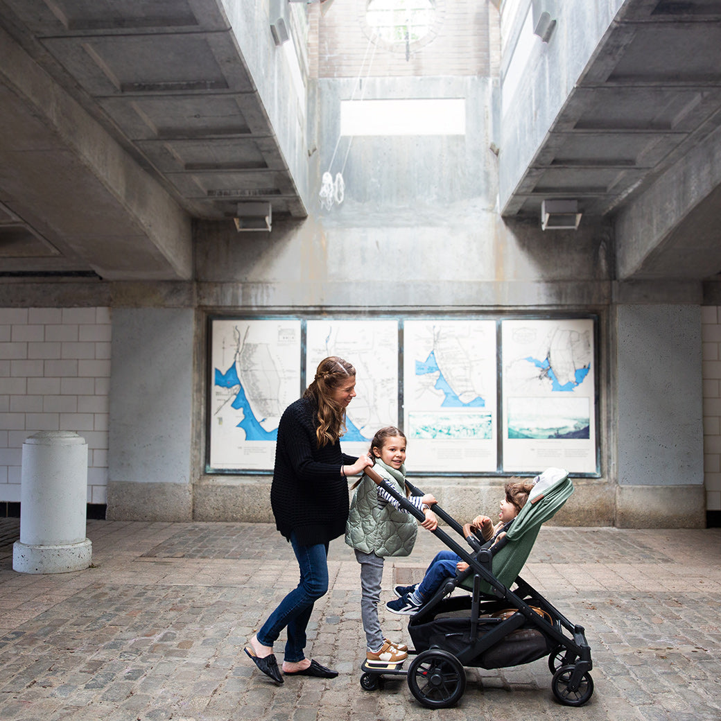 Woman pushing CRUZ V2 Stroller in color gwen with kid standing on the piggyback and another child sitting in stroller -- Color_Gwen