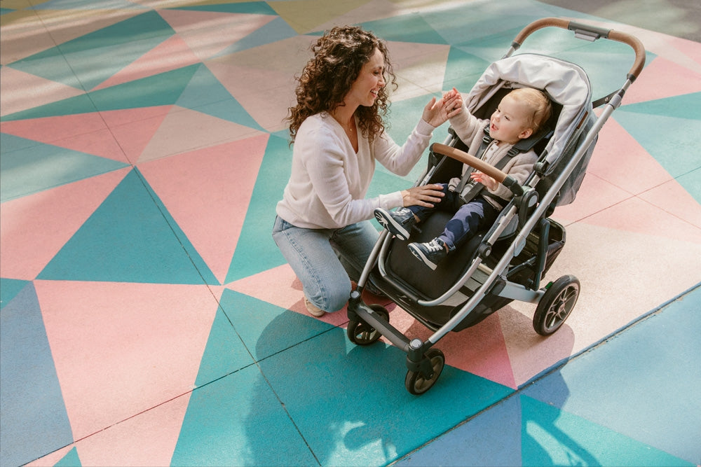Mother with curly hair wearing jeans and a loose fitting sweater, kneeling down to lovingly touch a smiling toddler seated in the uppababy cruz v2 stroller which is slightly angled towards the camera -- Lifestyle