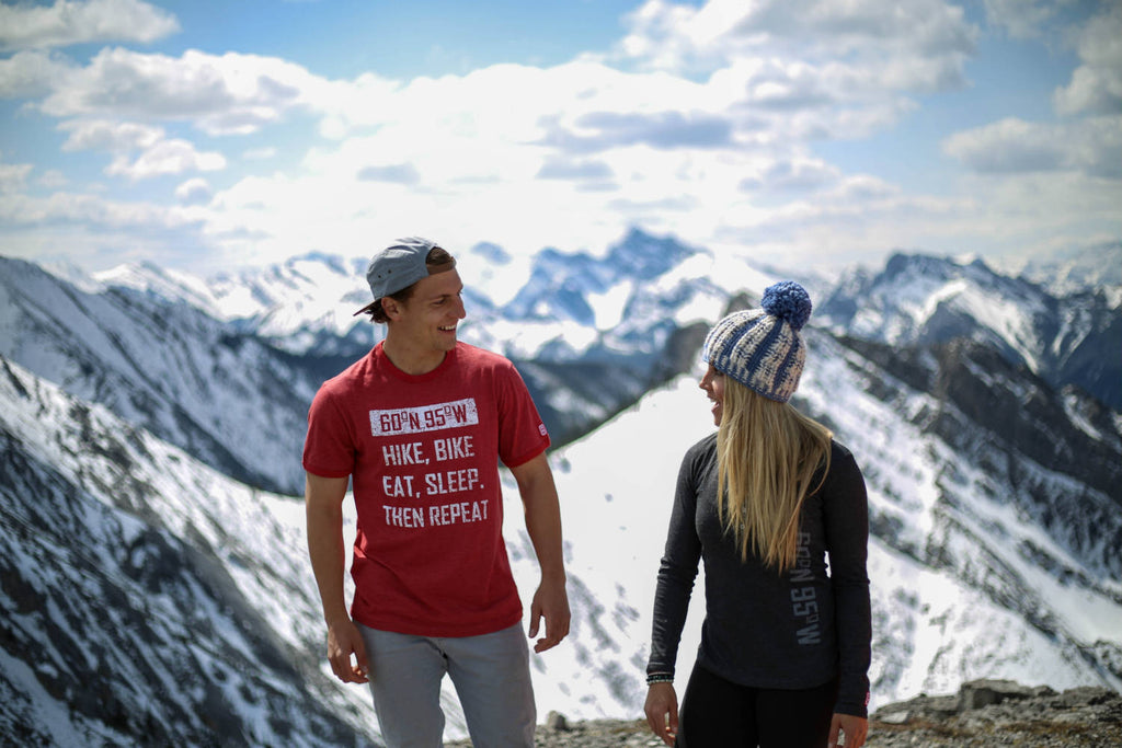 Girl and guy standing on top of a mountain wearing 60°N 95°W clothing smiling at each other