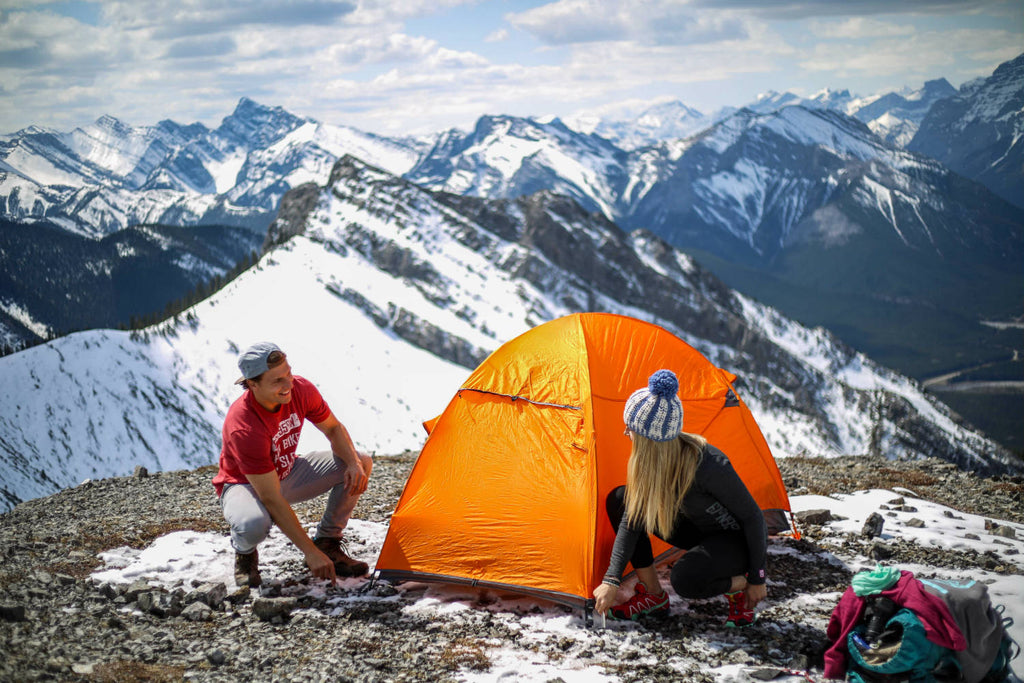 Girl and guy crouched down fastening their tent on top of a mountain