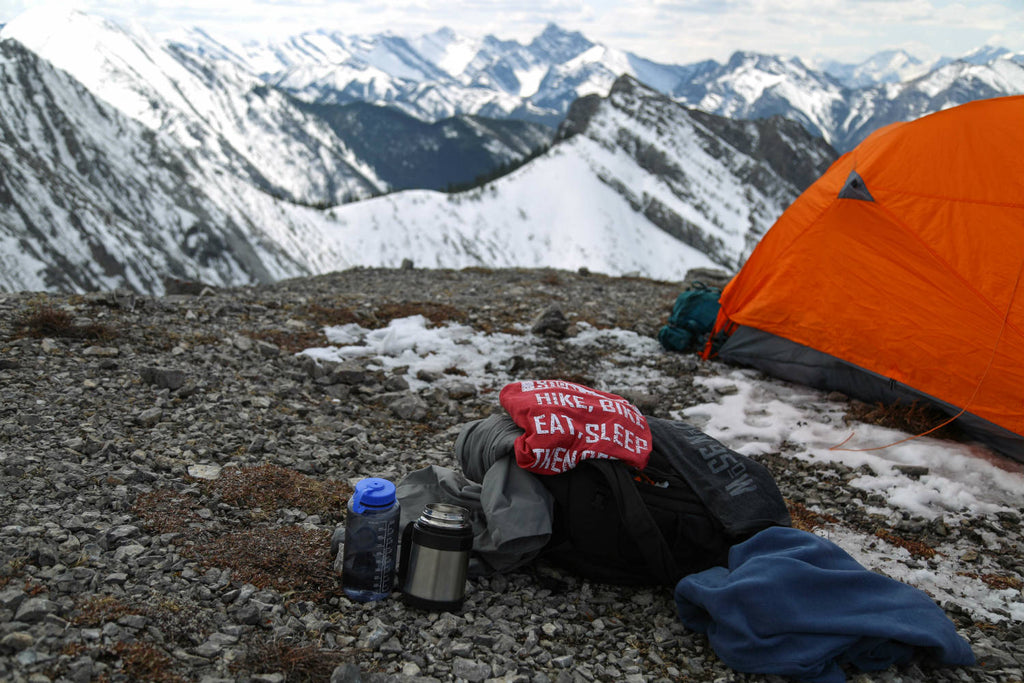 Camping gear on the ground with the 60°N 95°W faded red t-shirt and black mix 5-button henley