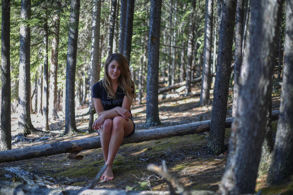 Girl wearing a 60°N 95°W black v-neck t-shirt sitting on a falled tree in the forest smiling at the camera