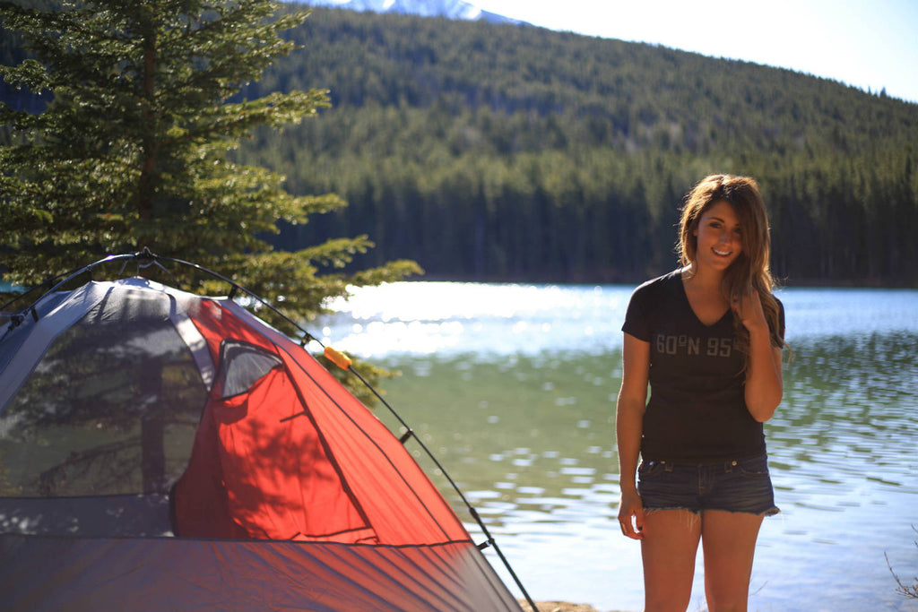 Girl standing by her tent by a lake wearing a 60°N 95°W black v-neck t-shirt