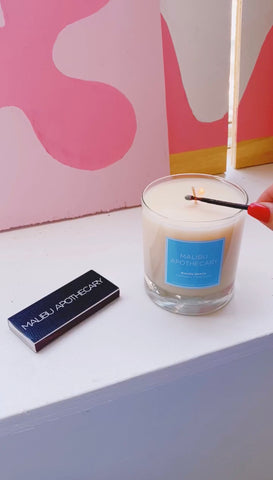 scented candle in a clear glass vessel with a blue label being lit with a match from Malibu Apothecary