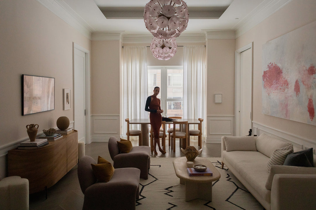 Designer Carly Cushnie stands in front of a wooden rectangular dining table with six modern wooden chairs with white upholstered seats. In front is a low modern sofa, oval wood coffee table, two low velvet armless chairs and a curved wooden sideboard.