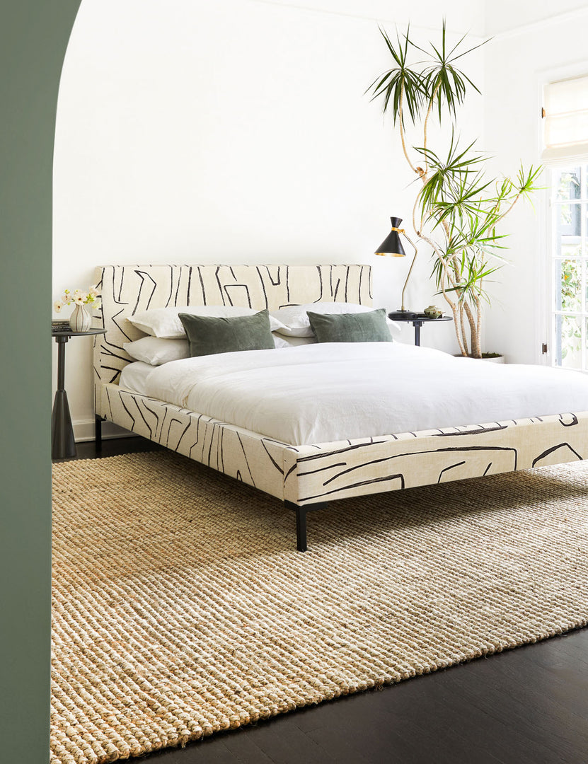 The Harriette scandinavian-inspired rug made of 100% jute lays in a bedroom with a cream and black patterend, framed bed, two black sculptural nightstands, and sage-toned velvet throw pillows