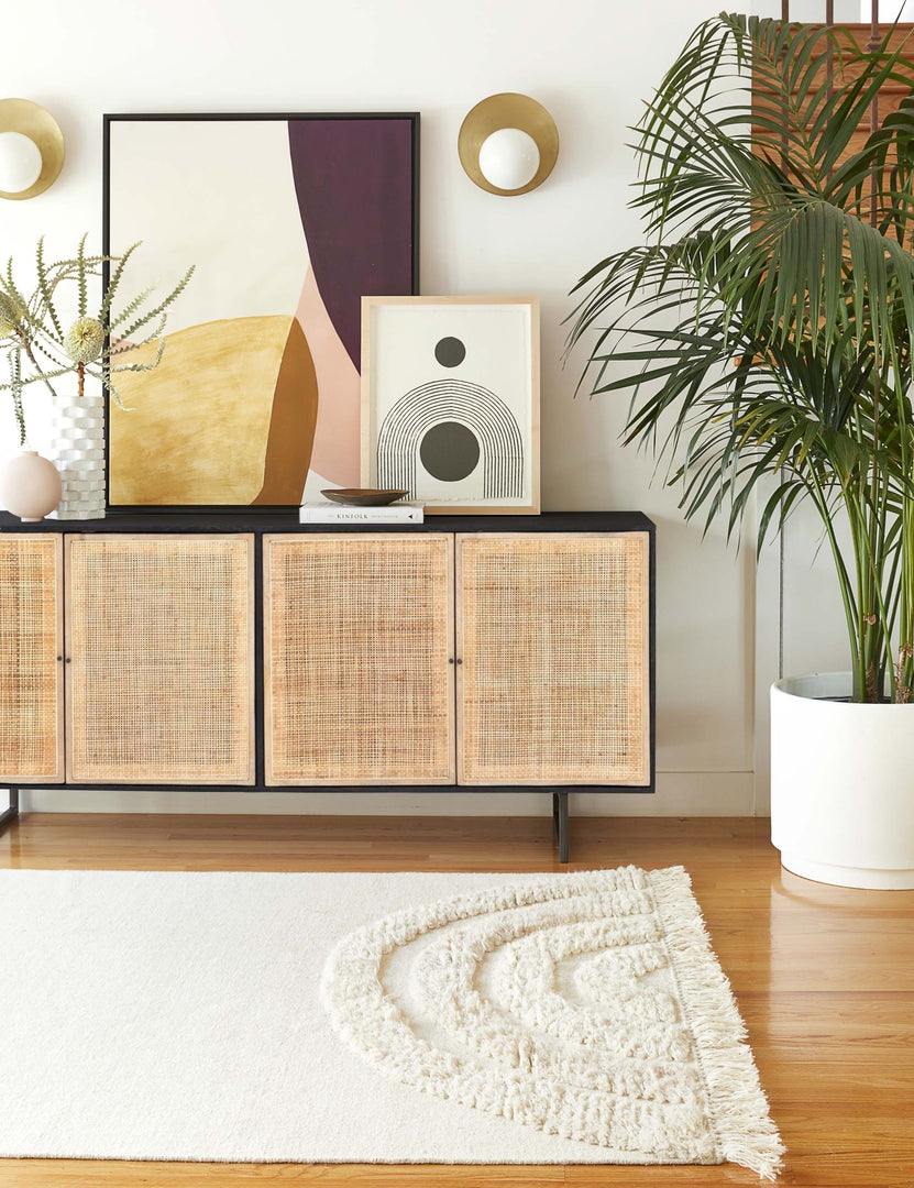 The Hannah black mango wood sideboard with cane doors and an iron base sits in a room with sculptural wall art sitting atop it and a plush white rug laying in front.