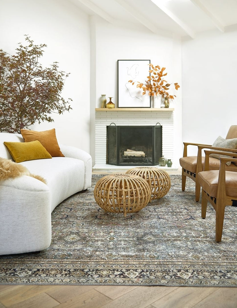 The Dacion distressed dark-toned persian rug lays in a living room with two jute ottomans, a rounded linen sofa, and two brown leather accent chairs