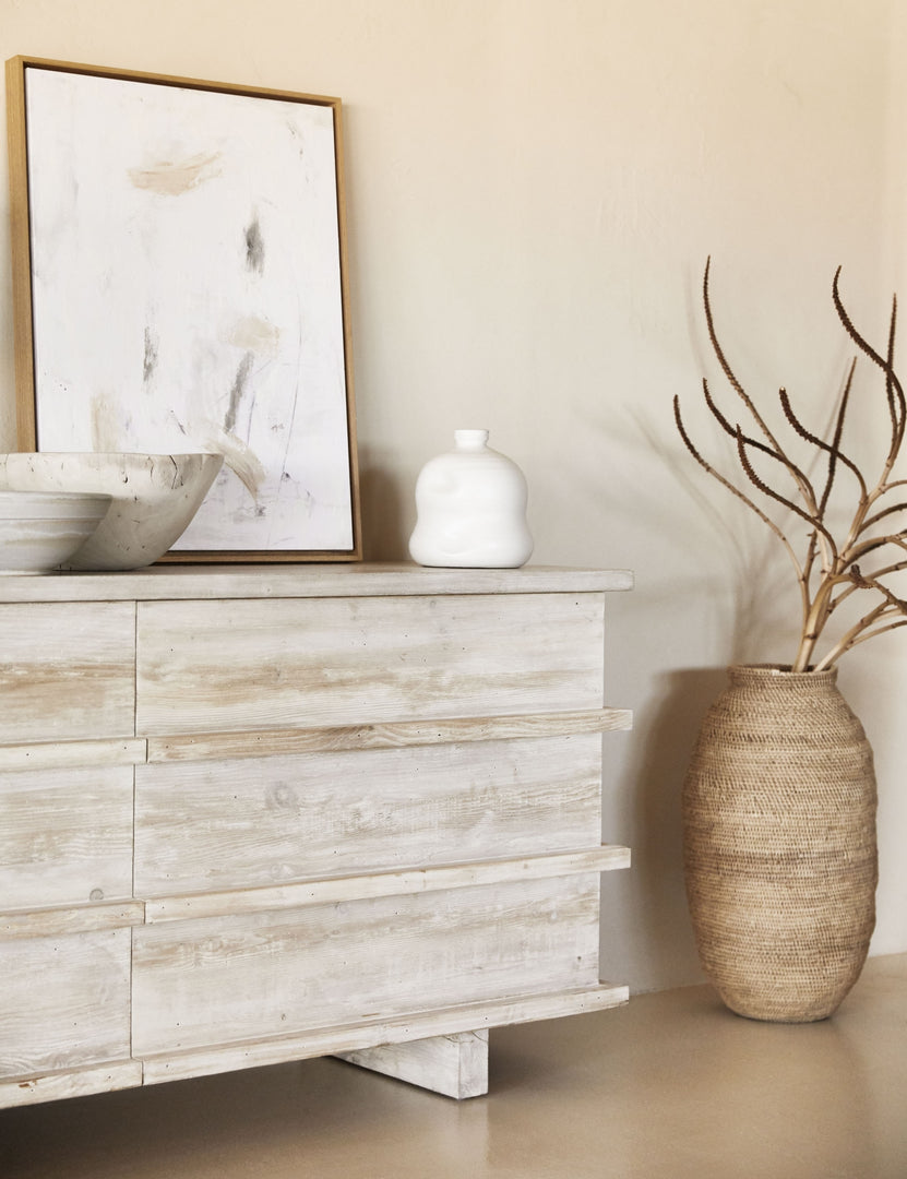 The Corliss 6-drawer white-washed wooden dresser has a white abstract painting leaning on top of it and decorative white bowls and vases.