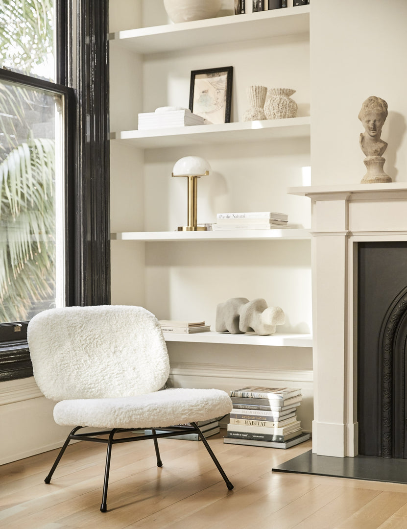 The Amanda white plush upholstered accent chair sits in the corner of a living room in front of a shelf with various stacked books and sculptural objects.