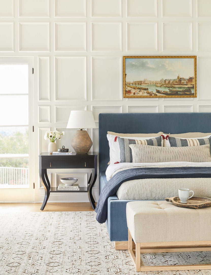 The Lockwood blue velvet-upholstered bed with a white oak base sits atop a patterned hand-woven rug against a white, accented wall.